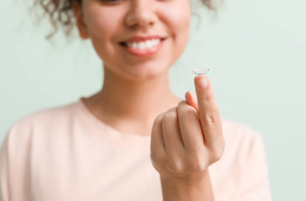 Close up image of a young smiling brunette woman's extended finger with a contact lens on it against a light green background.