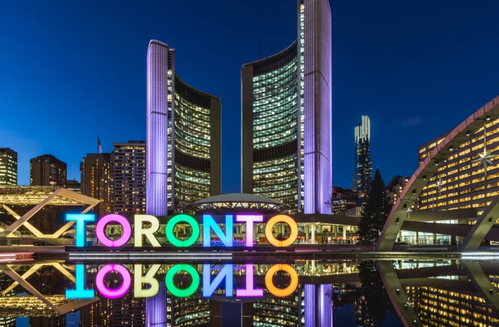 Image of the Toronto sign and city hall at Nathan Philips square lit up in colours during the evening at blue hour.