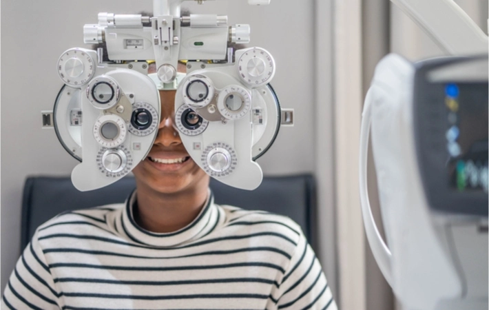 Young smiling female patient being assessed with a phoropter for her eye exam and an optometrist clinic.
