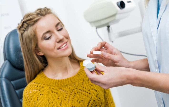 Young brunette woman sits in an examination chair at the optometrist's office as the eye doctor shows her a contact lens case and its contents.