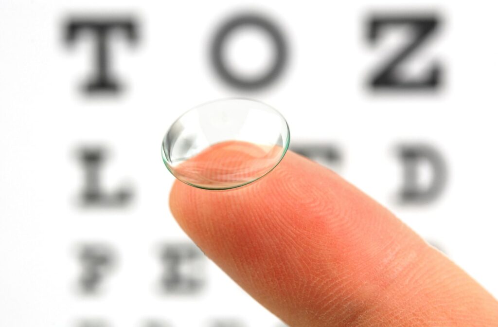 A close-up image of a contact lens sitting on someones index finger, with a Snellen eye chart blurred in the background
