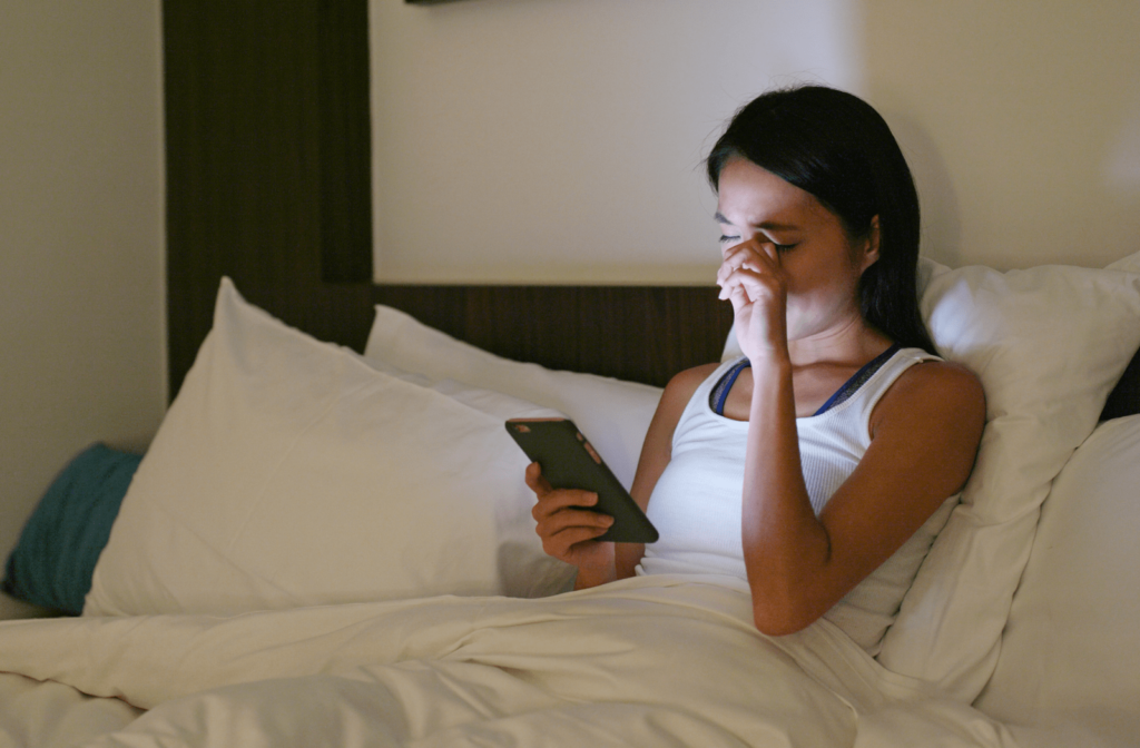A young woman lying in bed, looking at her cell phone and rubbing her eye