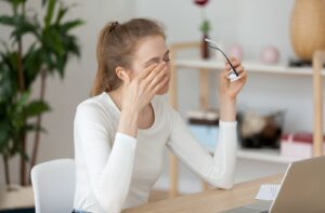 A young woman sitting at the desk at work had removed her eyeglasses and is massaging her eyes due to vision blurriness