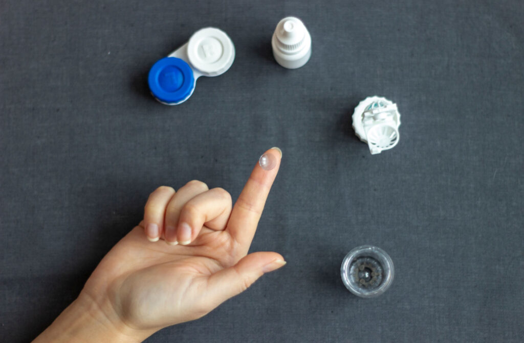 Bird's-eye view of a person's right hand with a contact lens on their index finger above a table with a contact lens case, contact solution, and eye drops on it