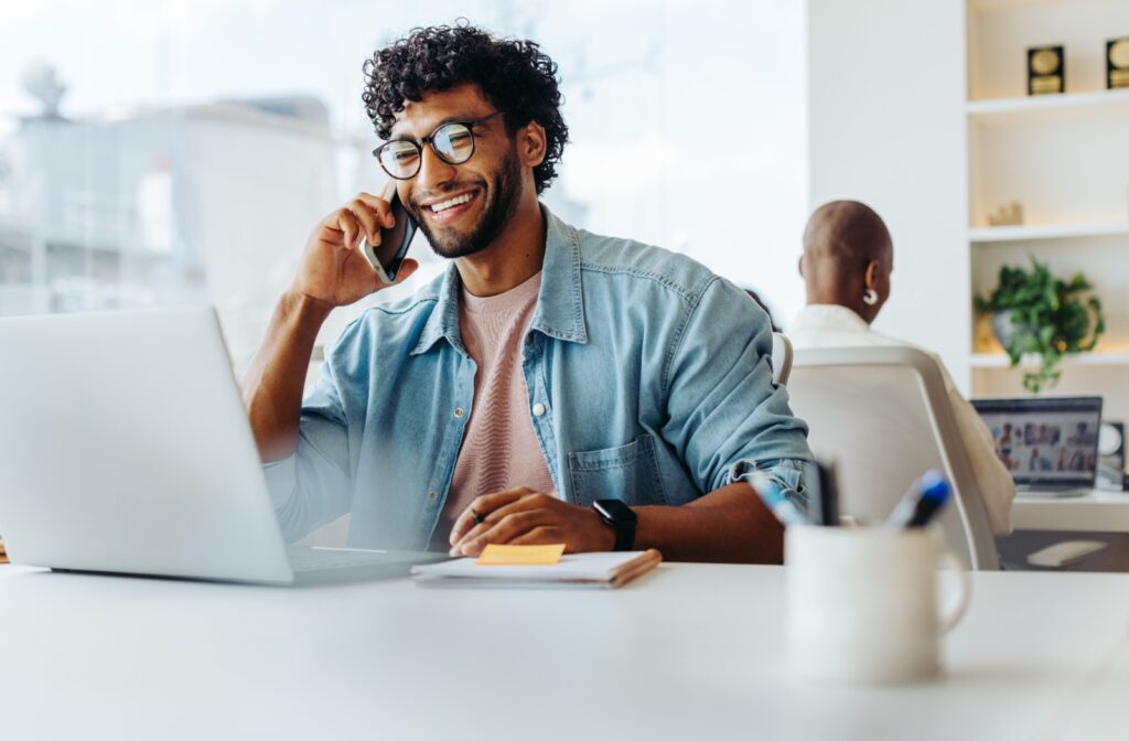 A businessman wearing glasses smiles while on the phone and using a laptop while in a brightly lit office space