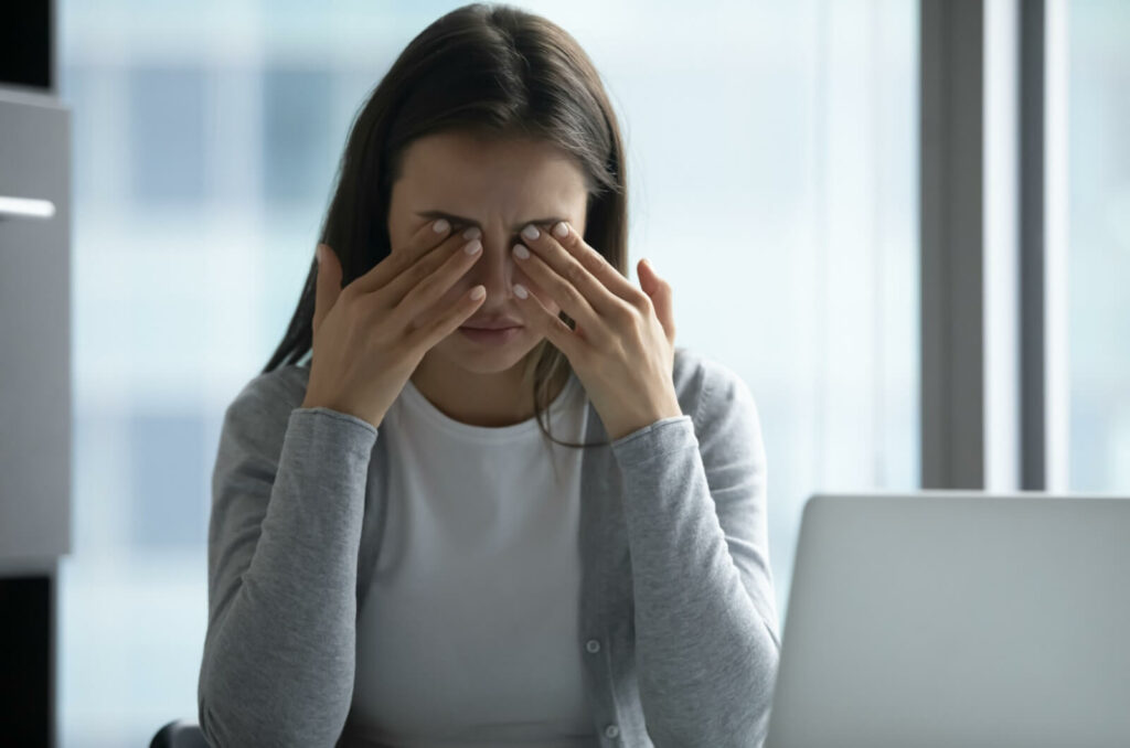 A young adult at their computer, rubbing their eyes in discomfort due to their dry eye discomfort.