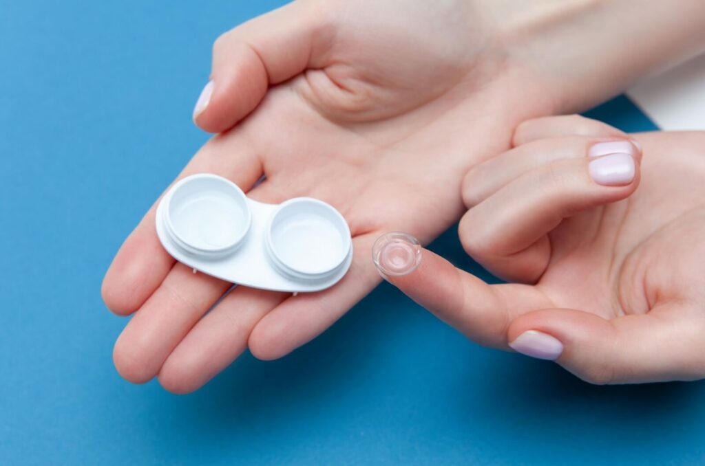 A scleral contact lens on the tip of a person's finger with a white case resting in their hand over a blue background.