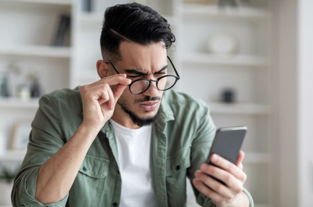 A young adult adjusting their glasses and squinting in frustration trying to read their phone screen while adjusting to new glasses.